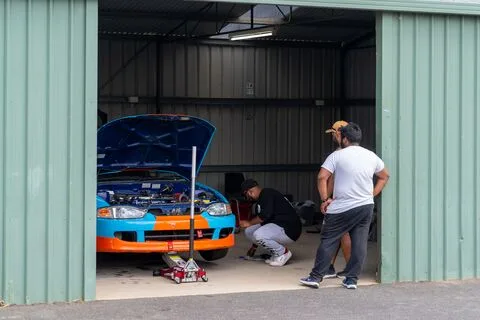 Two people working on a light blue and orange hatchback in a garage with the bonnet up