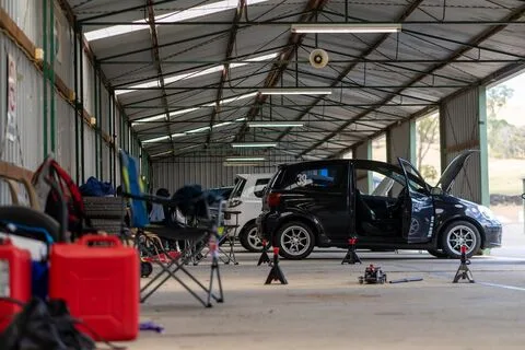 A black hatchback on jack stands in an open shed with fuel cans and folding chairs