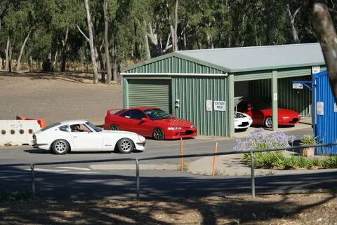 A white classic coupe and two red cars beside a green shed
