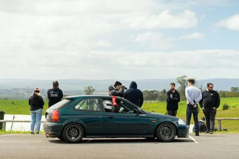 People standing trackside beside a dark green hatchback with a helmeted driver