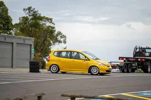 A yellow hatchback parked by the garages next to a car trailer