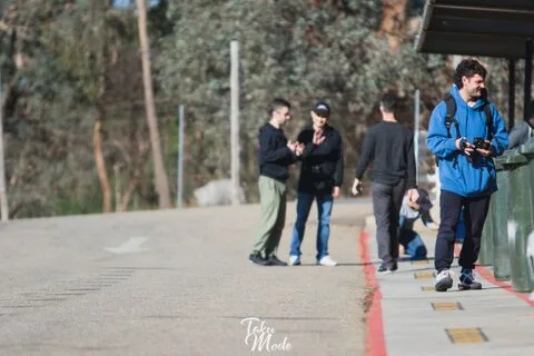 People chatting on a path beside a fence, one holding a camera
