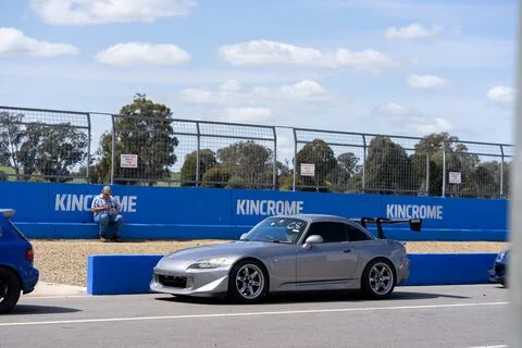 A silver coupe with a rear wing passing a blue Kincrome barrier as a spectator sits on the wall